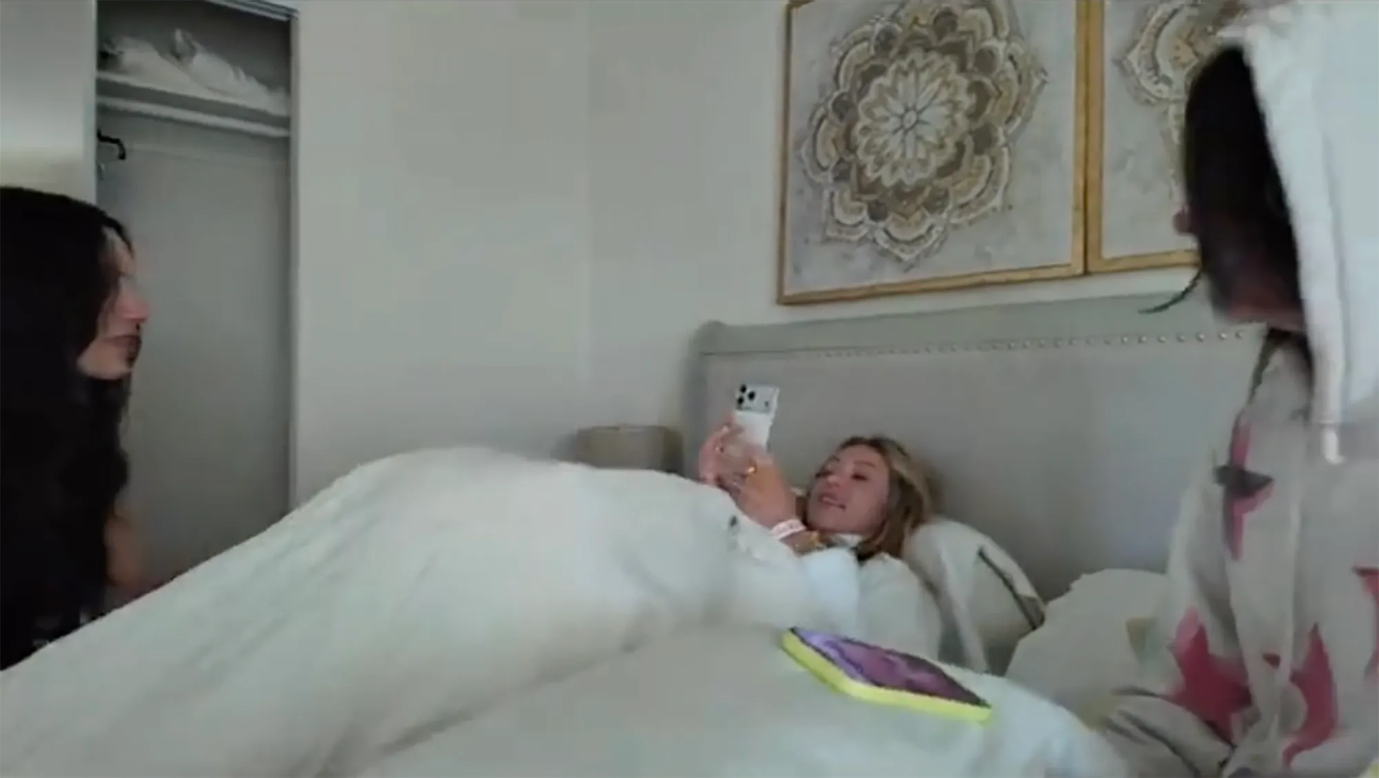 Alix Earle in bed, looking at her phone, with two other women beside her.