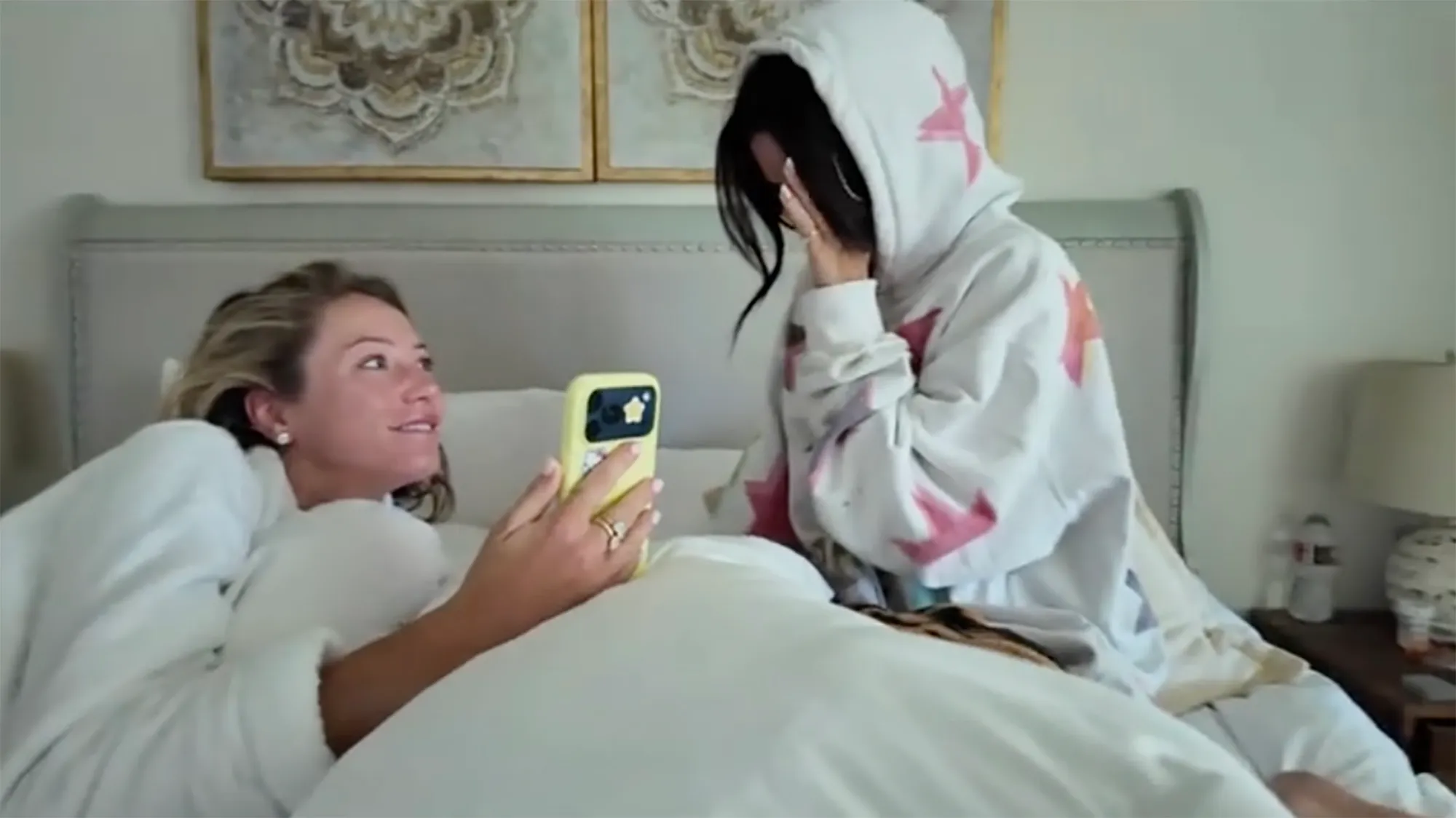 Alix Earle, in a white and pink star hoodie, covers her face as she reacts to a video on a phone held by another woman.