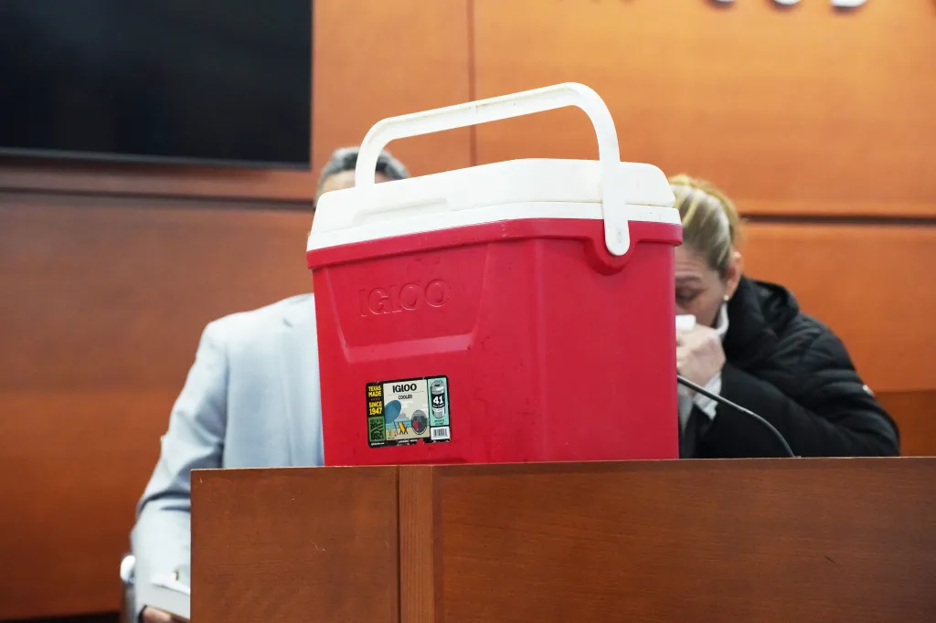 A red and white Igloo water cooler sits on a wooden barrier in a courtroom with people in the background.