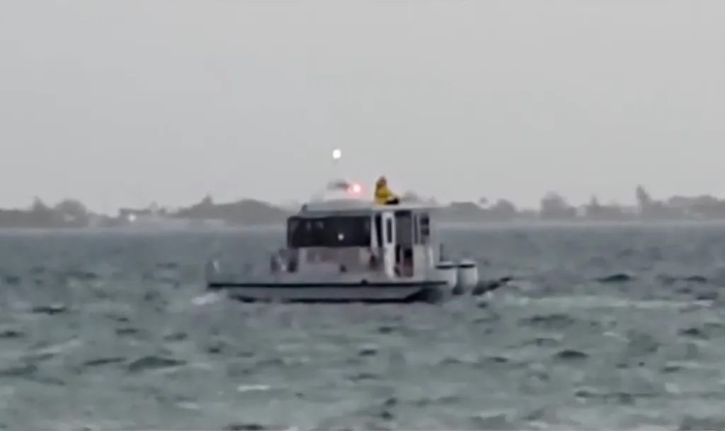 A small boat with a person in a yellow jacket on top, on rough water, with a blurry shoreline in the distance under a grey sky.