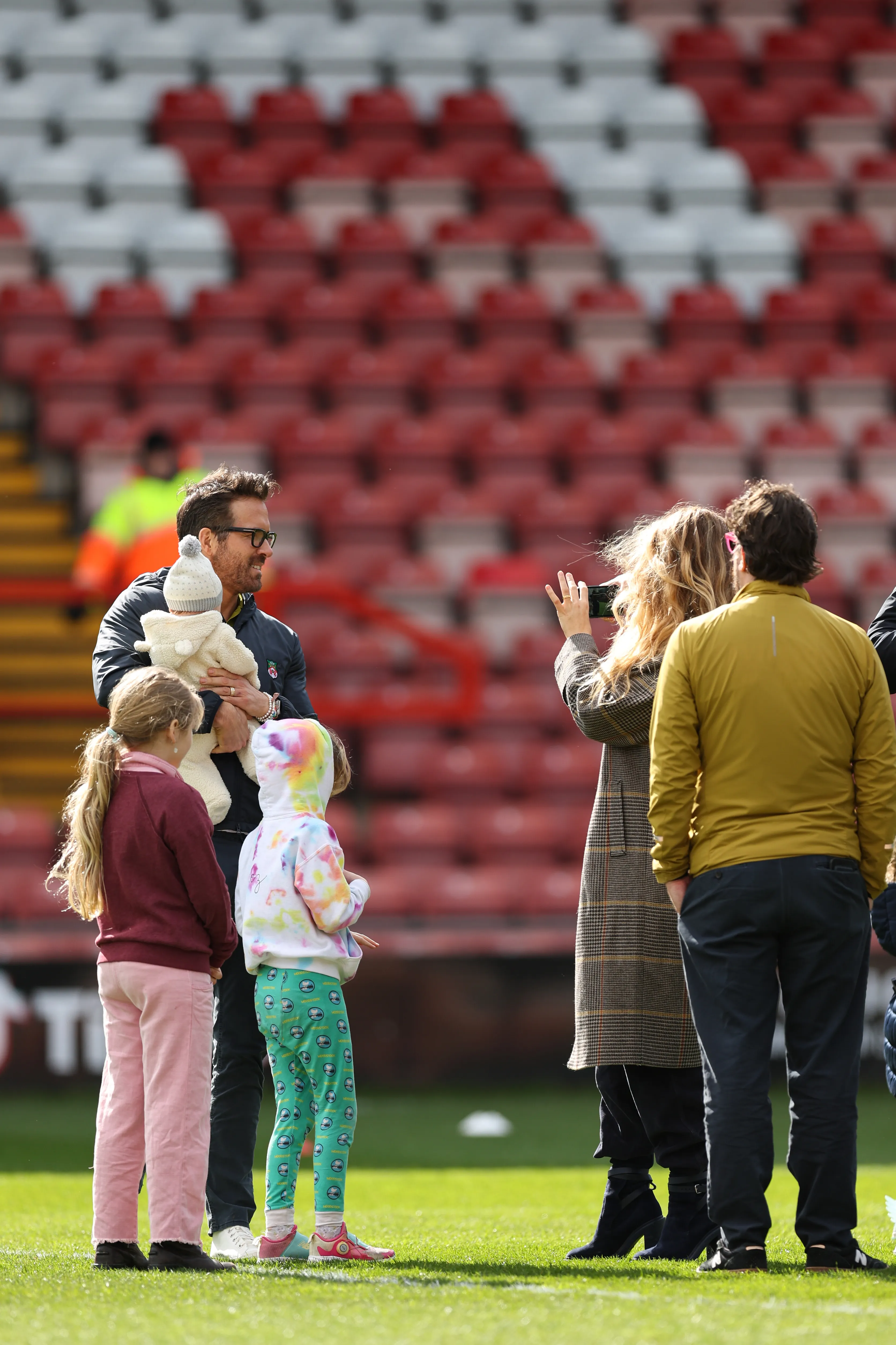 Ryan Reynolds holding a baby, with Blake Lively taking a photo, and their children on the field at The Racecourse Ground.