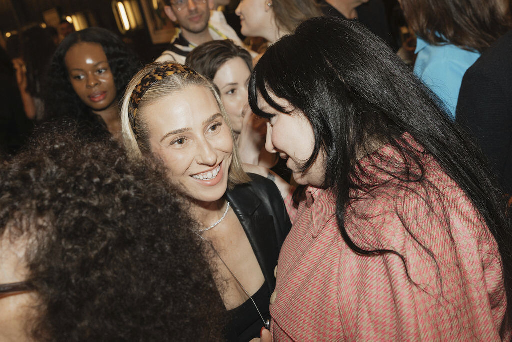 Lena Dunham smiling and leaning towards a woman in a crowded room.