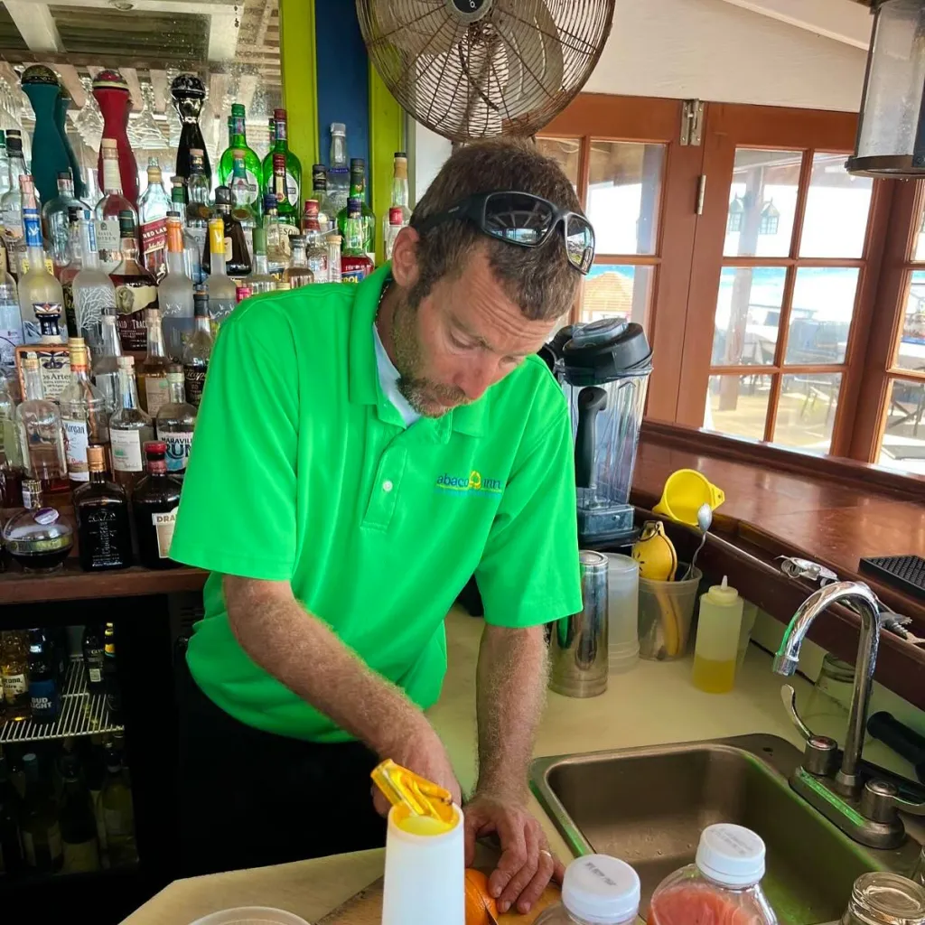 A bartender in a green "Abaco Inn" shirt preparing a drink ingredient.