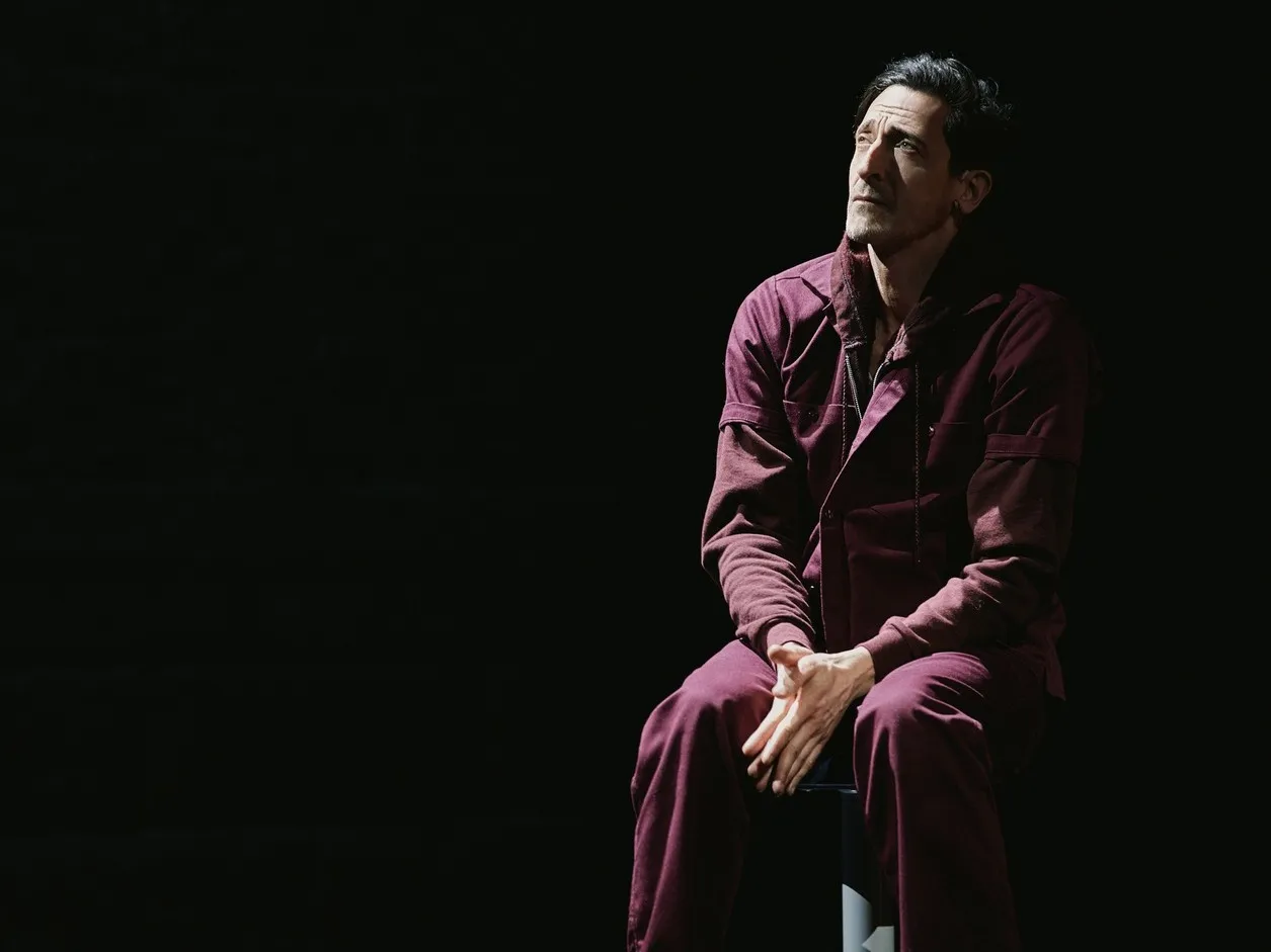 Adrien Brody in a maroon jumpsuit looking up while sitting.