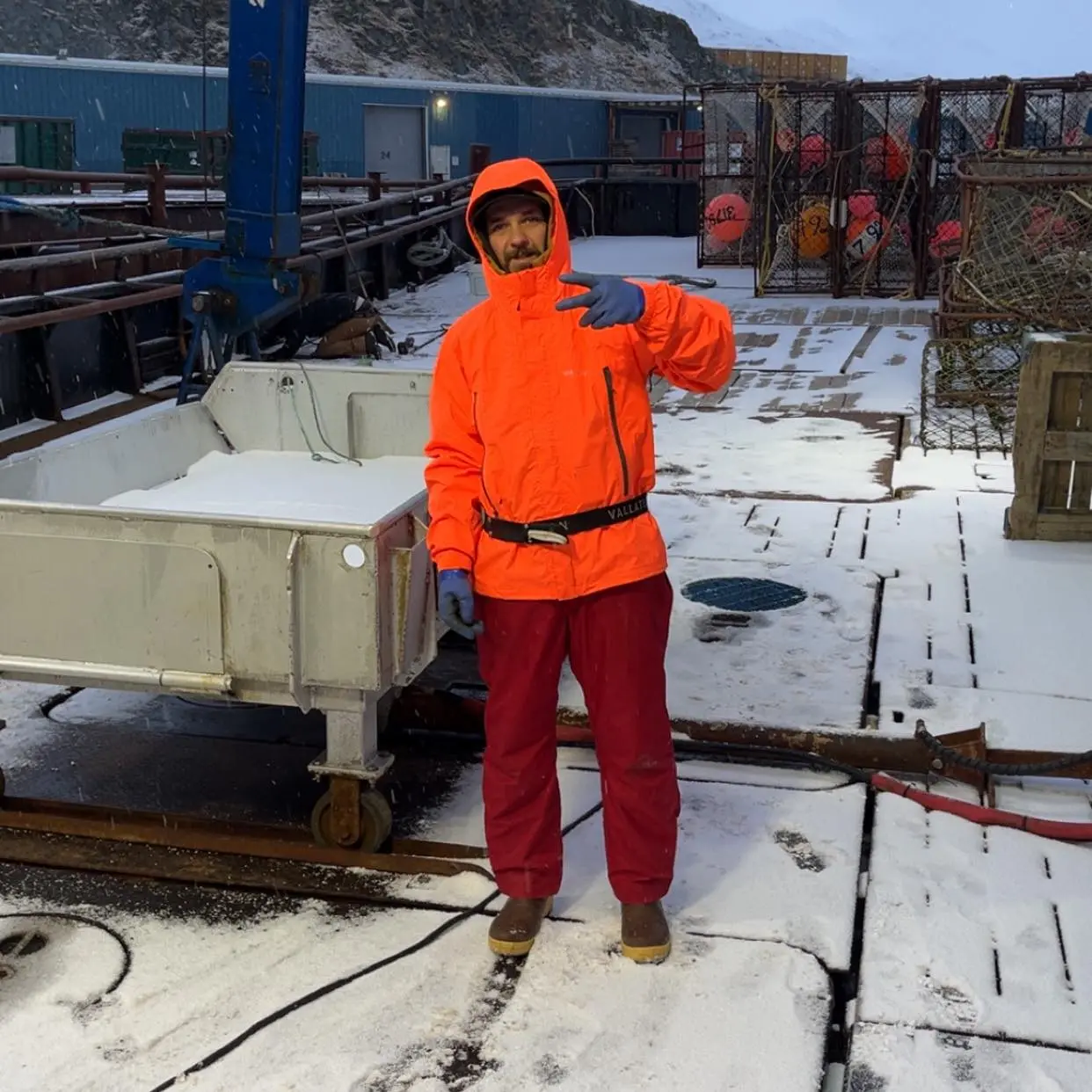 Todd Meadows standing on a snow-covered boat deck, wearing an orange jacket and red pants, making a peace sign with his left hand.