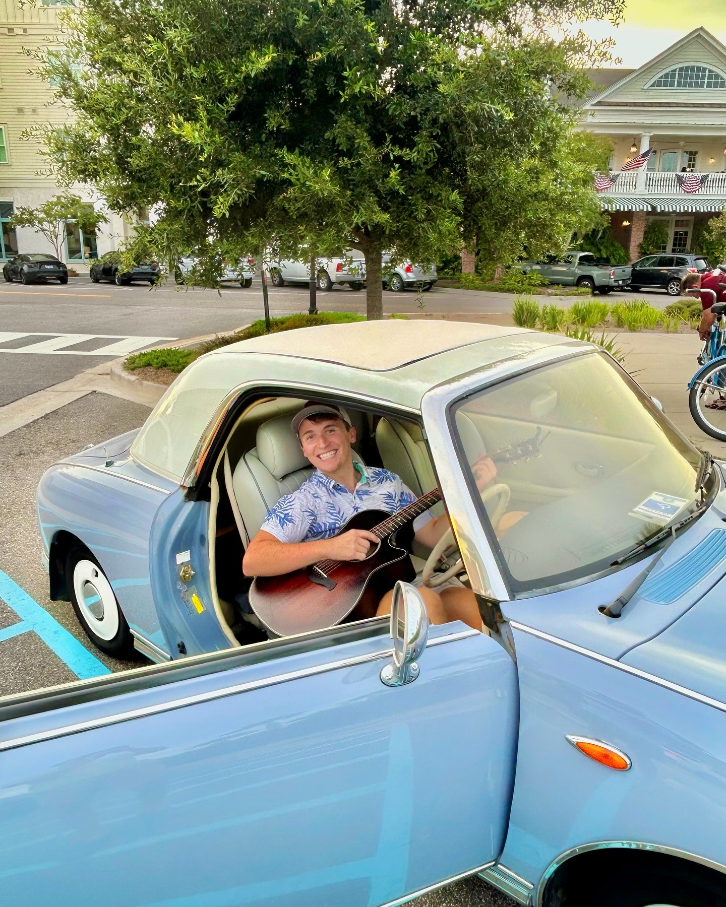 Dylan Carter in a light blue convertible, playing an acoustic guitar and smiling.