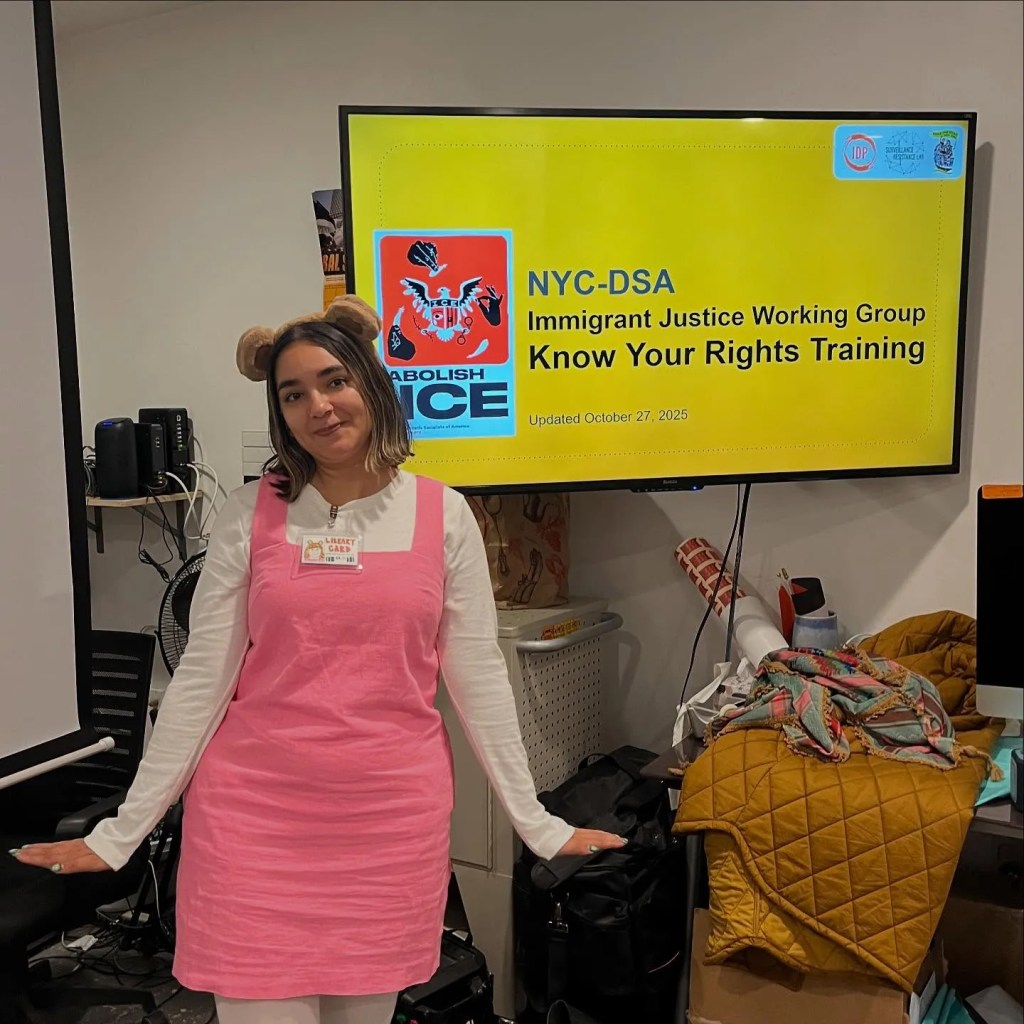 A woman in a pink dress and a bear-ear headband stands in front of a monitor displaying "NYC-DSA Immigrant Justice Working Group Know Your Rights Training."