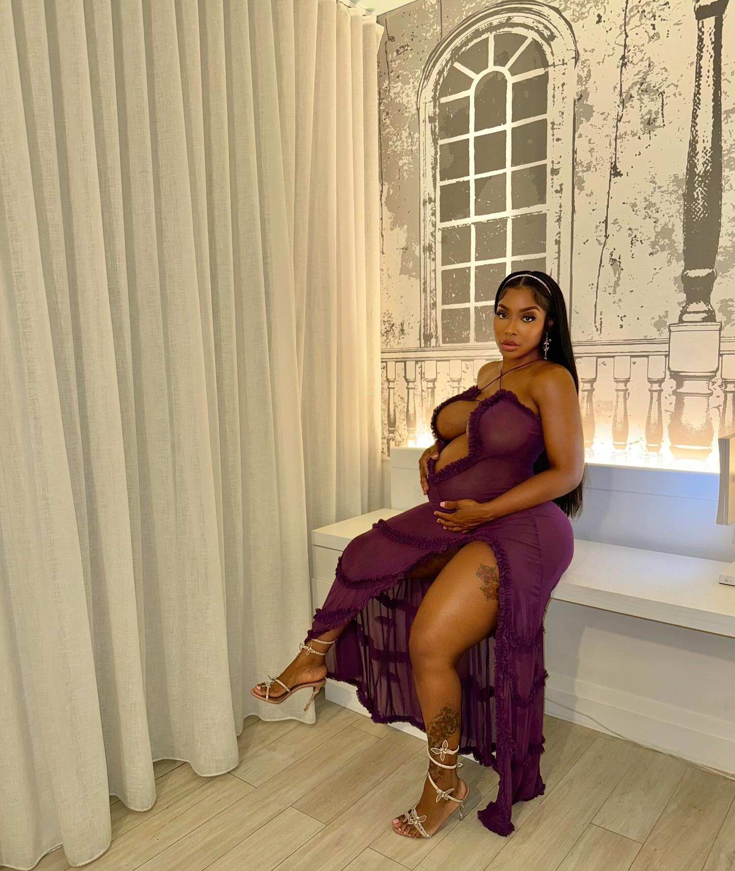 Ayesha Howard, a woman with long dark hair, wearing a flowing sheer purple dress and strappy high-heeled sandals, sits on a white bench.