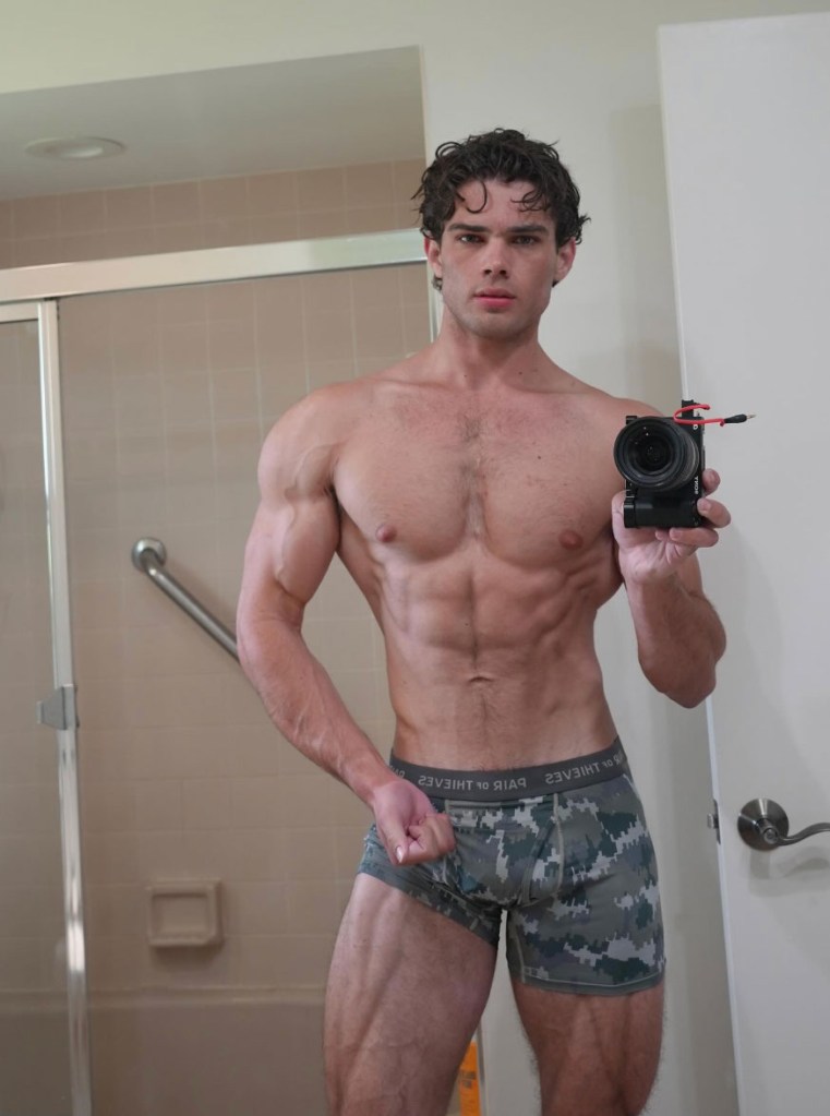 A muscular man with curly hair taking a shirtless mirror selfie in a bathroom while holding a camera.