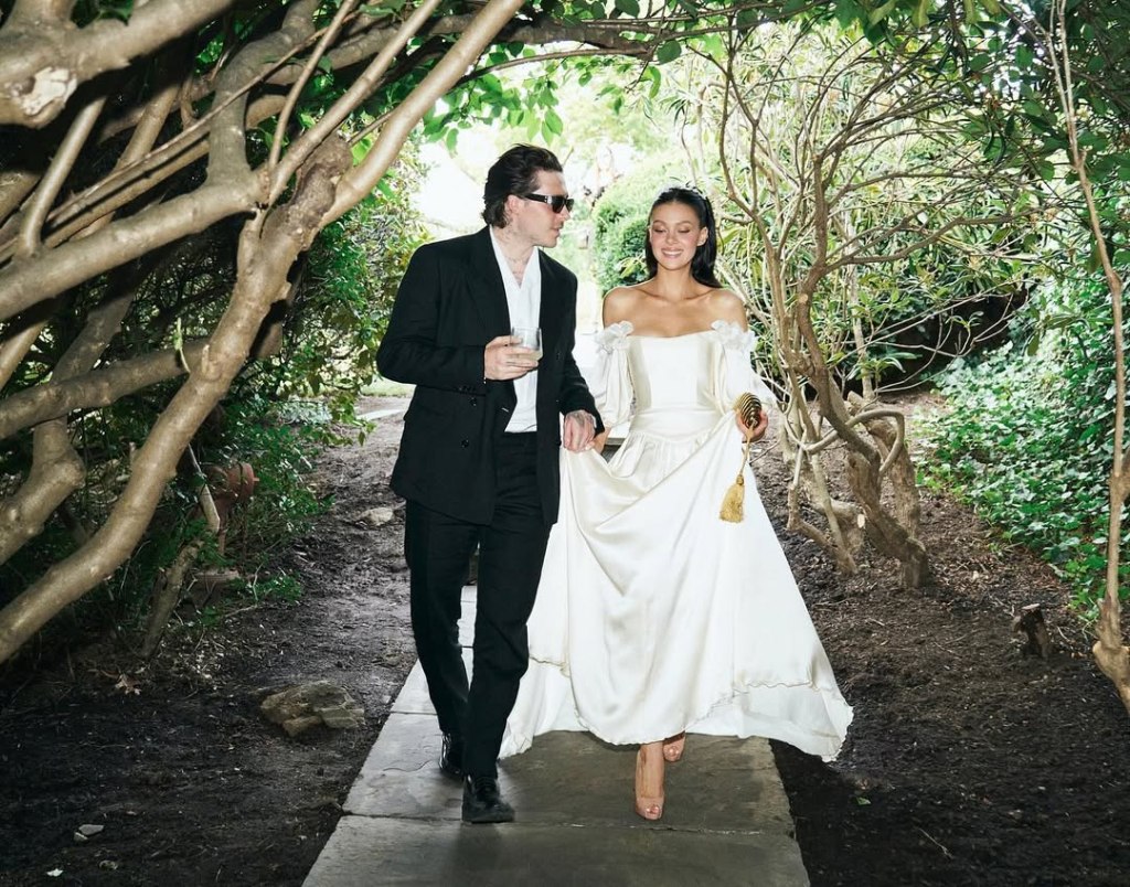 Brooklyn Beckham, in a black suit and white shirt and sunglasses, and Nicola Peltz, in an off-the-shoulder cream wedding gown, walking together, with Peltz holding her wedding dress.