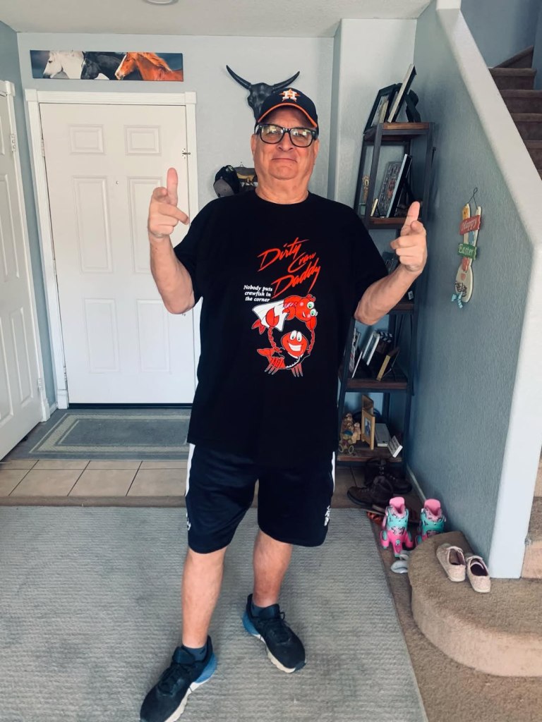 A man in a black T-shirt and shorts, wearing an Astros baseball cap and glasses, points at the camera.