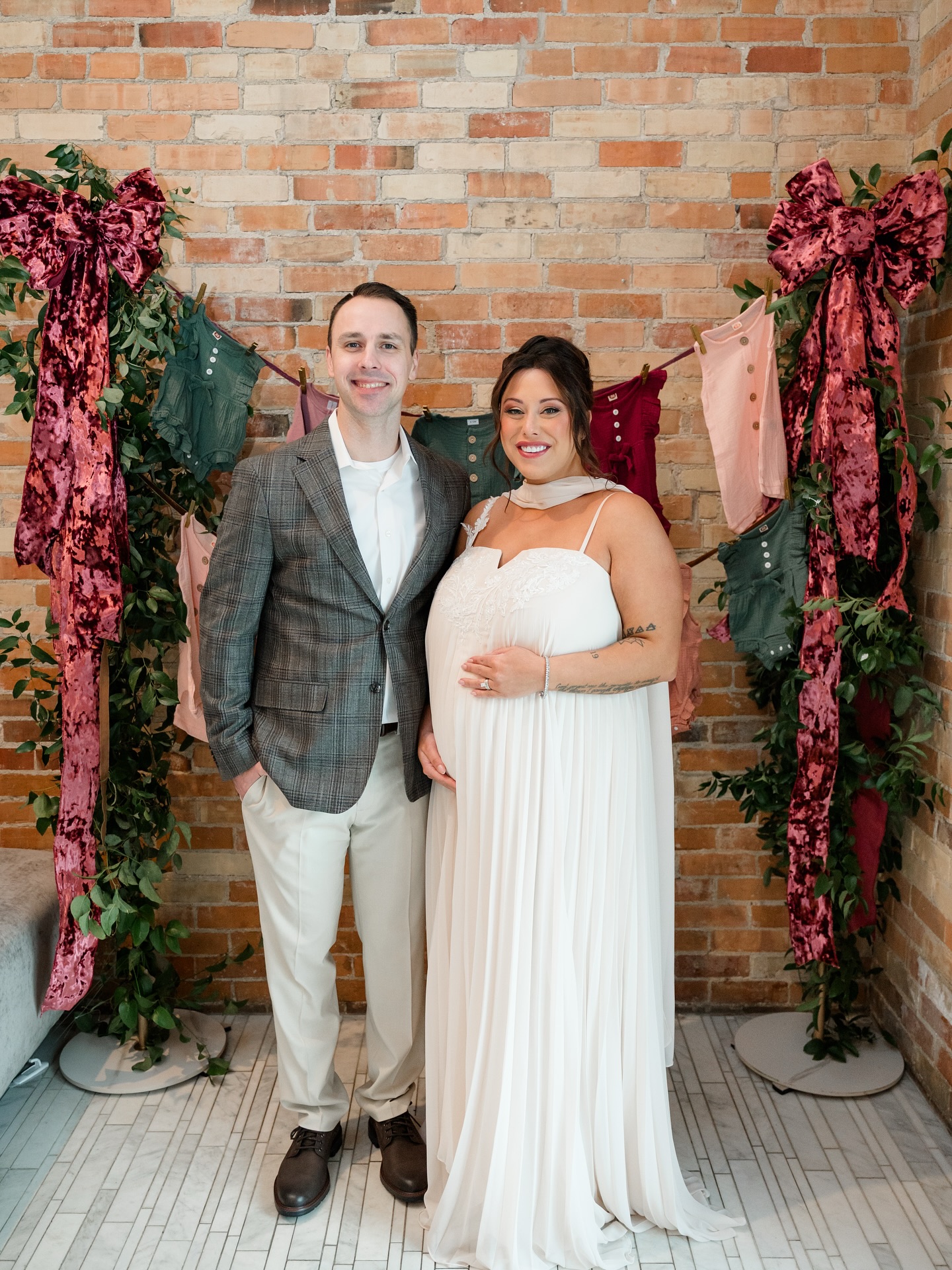 A couple posing for a photo against a brick wall, with the pregnant woman in a white dress cradling her belly.