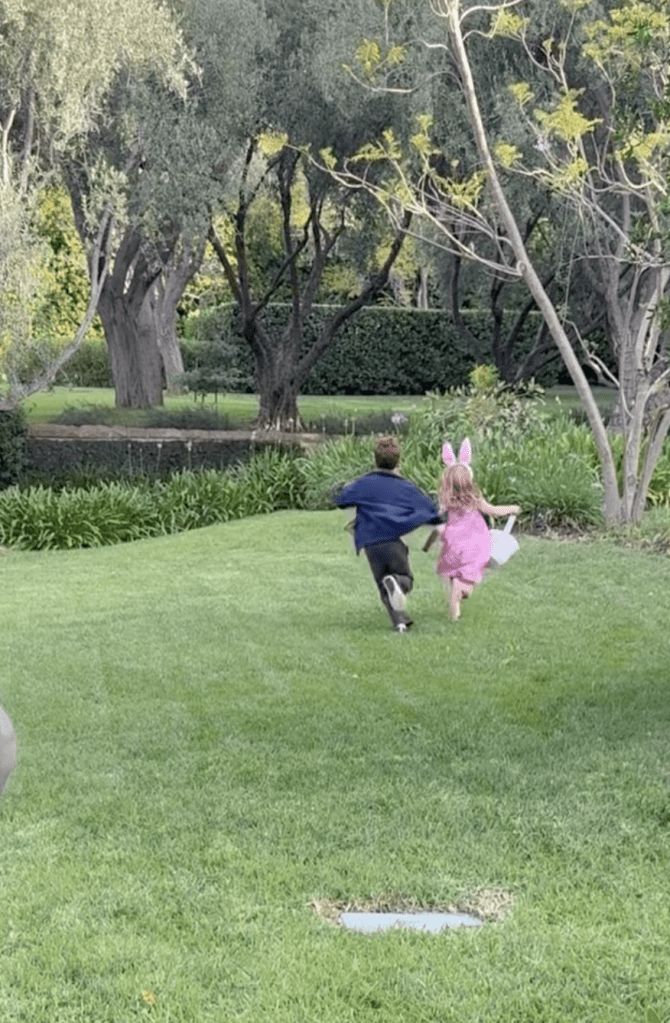 Prince Archie, in a blue jacket and black pants, runs with Princess Lilibet, in a pink dress and pink bunny ears.