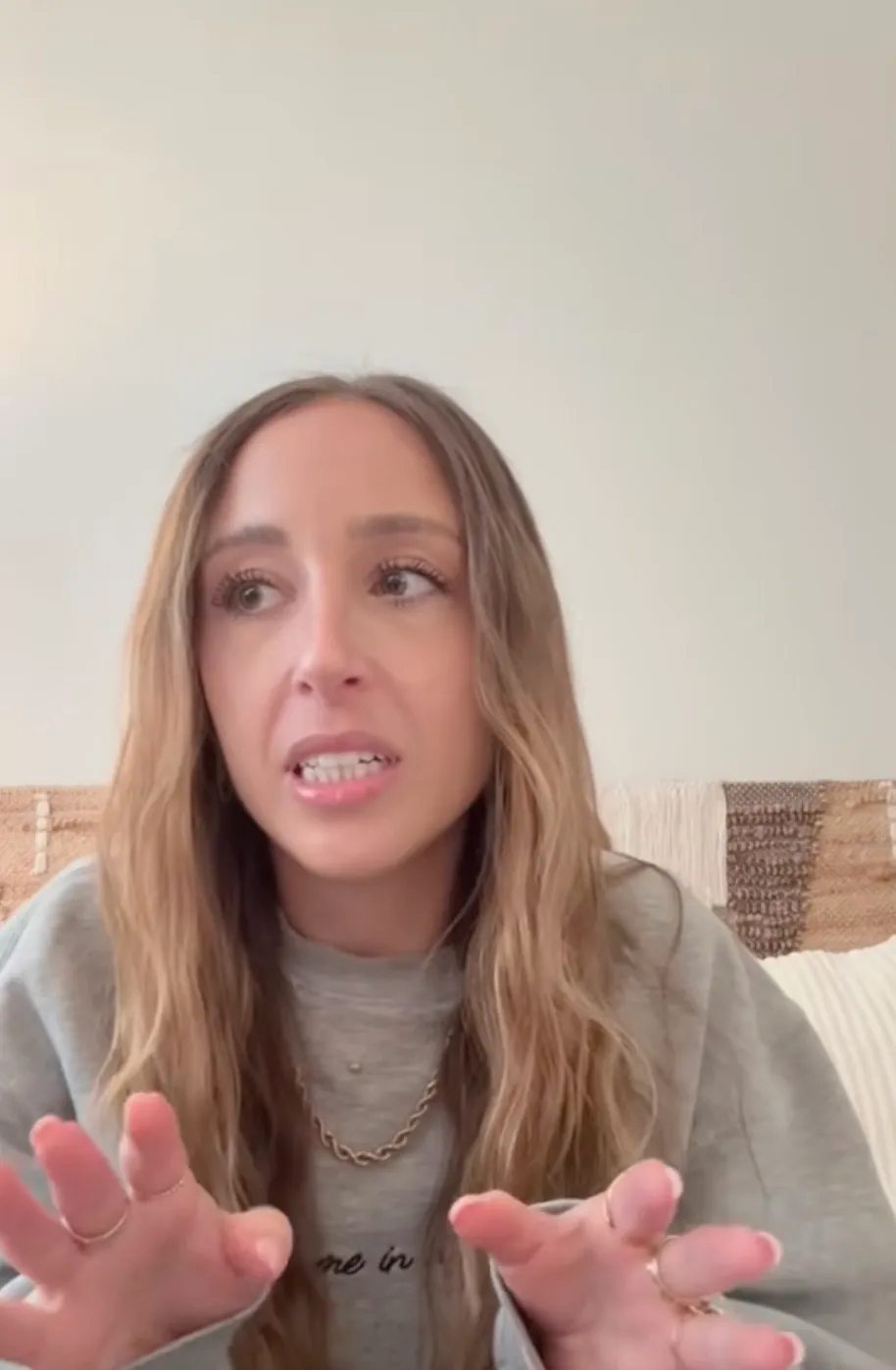 A woman with long brown hair, wearing a gray sweatshirt and a gold chain, gestures with her hands while speaking.