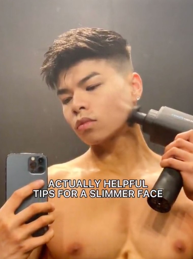 A man using a massage gun on his jawline while holding a phone in a mirror with the text "ACTUALLY HELPFUL TIPS FOR A SLIMMER FACE".