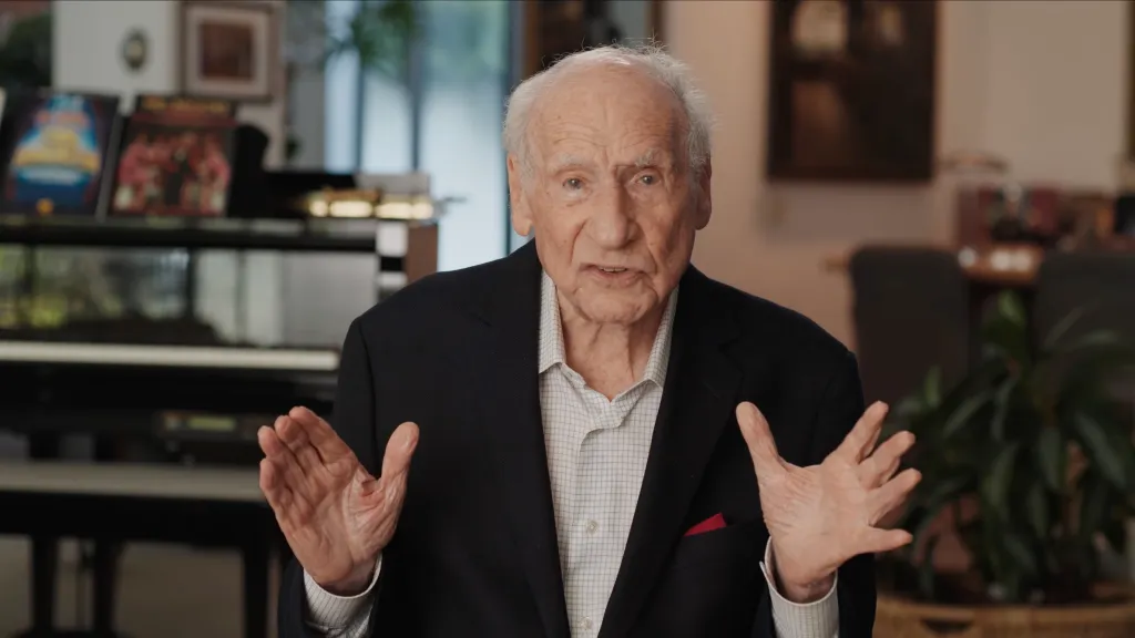 Mel Brooks speaking to the camera, gesturing with both hands.