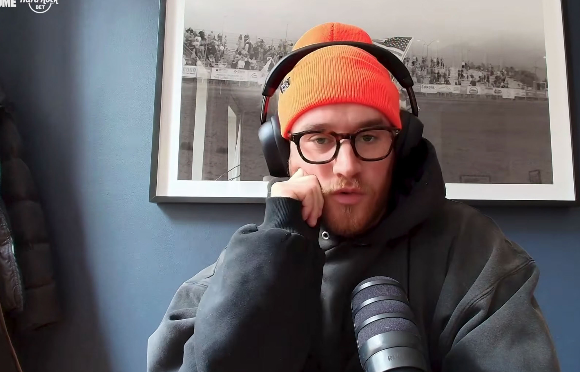 A man in an orange beanie, headphones, and glasses speaking into a microphone.