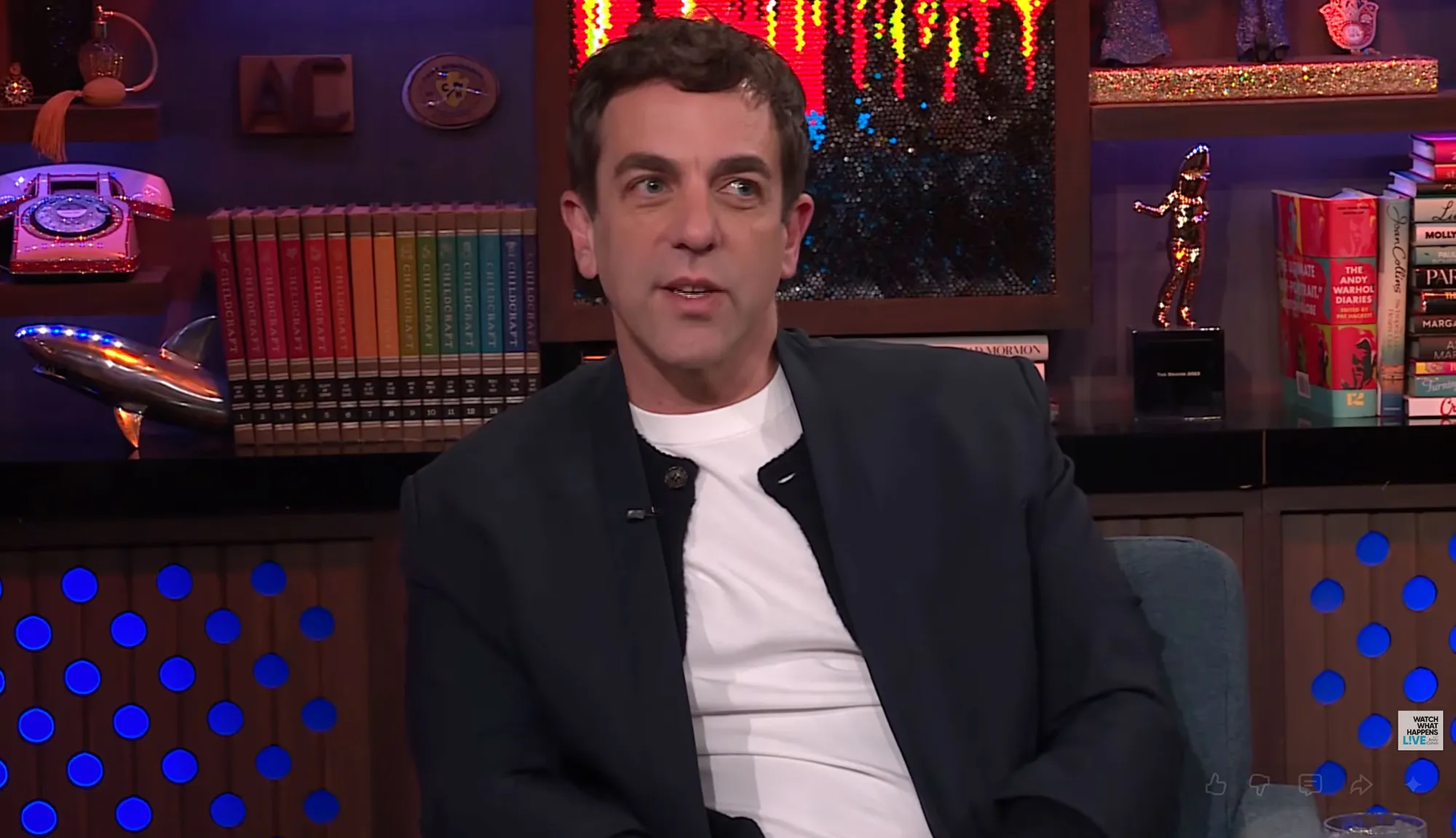 BJ Novak sitting on a blue couch, wearing a white shirt under a black jacket.