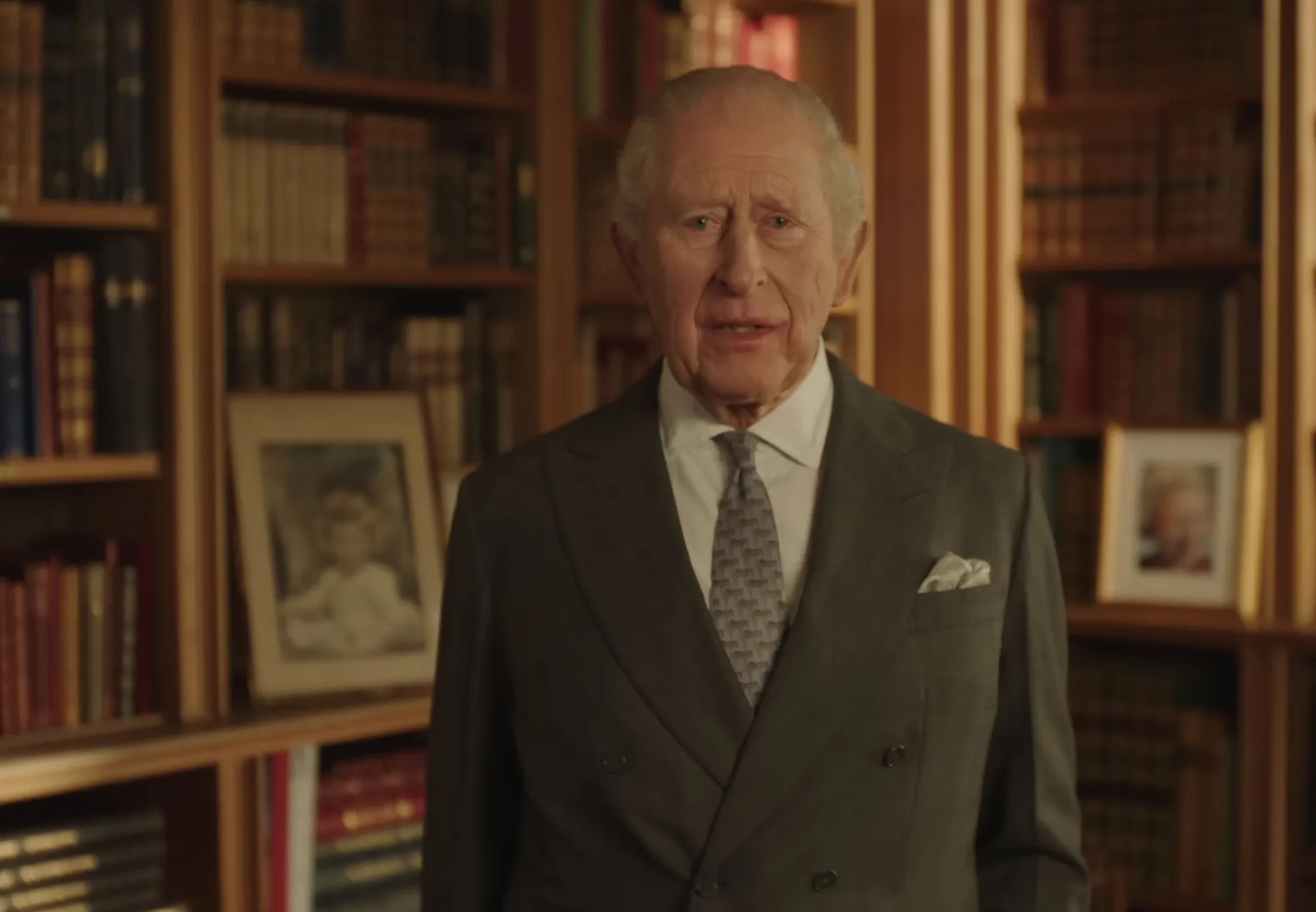 King Charles III in a grey suit and patterned tie, standing in front of bookshelves with framed photos.