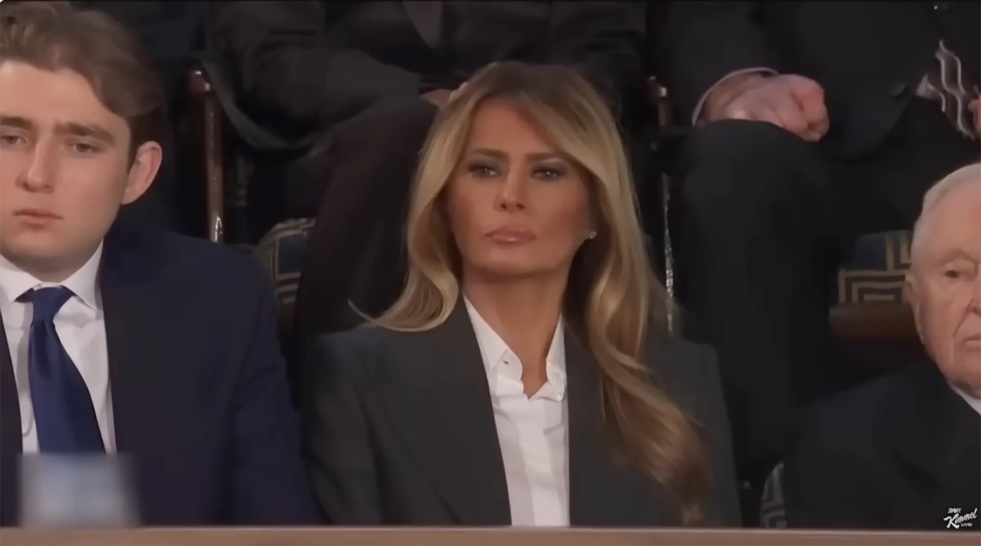 Melania Trump looking forward, seated between a younger man and an older man.