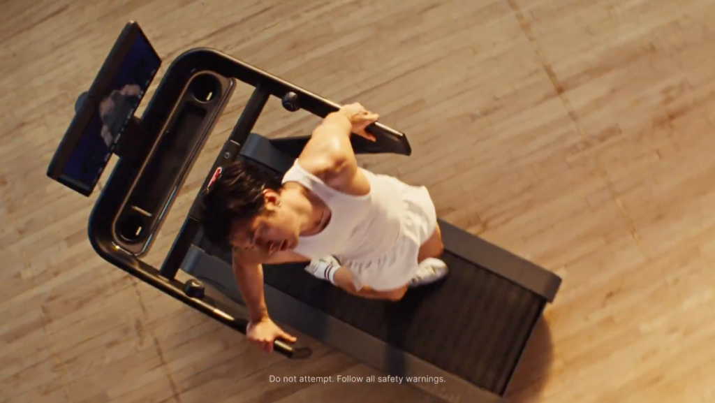 A man in a white tank top and shorts runs on a Peloton treadmill viewed from above.