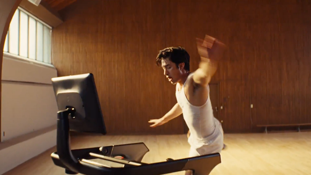 A man in a tank top runs on a Peloton treadmill in a wood-paneled room.