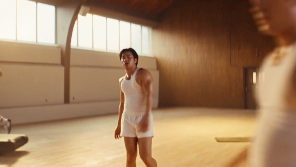 A young man in a white tank top and shorts stands in a well-lit gym with another blurred figure in the foreground.