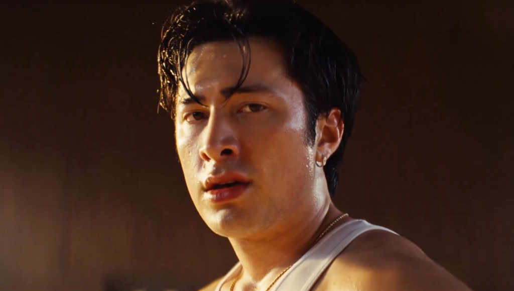 Close-up of a sweating man in a white tank top and gold chain looking intensely at the camera.