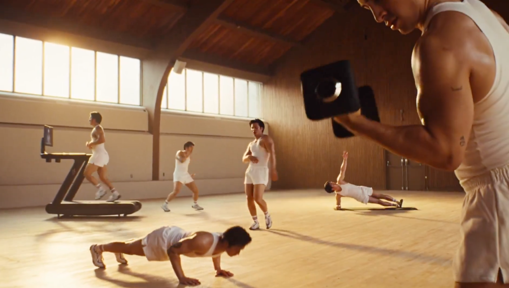 Multiple men working out in an airy room with large windows, some lifting weights, some doing pushups, one on a treadmill.