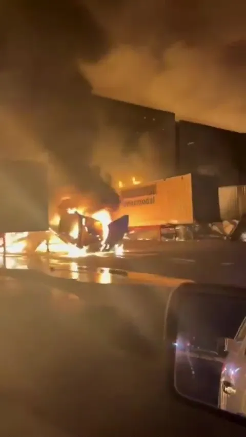 A burning truck and trailer with a large fire, smoke, and debris on the ground.
