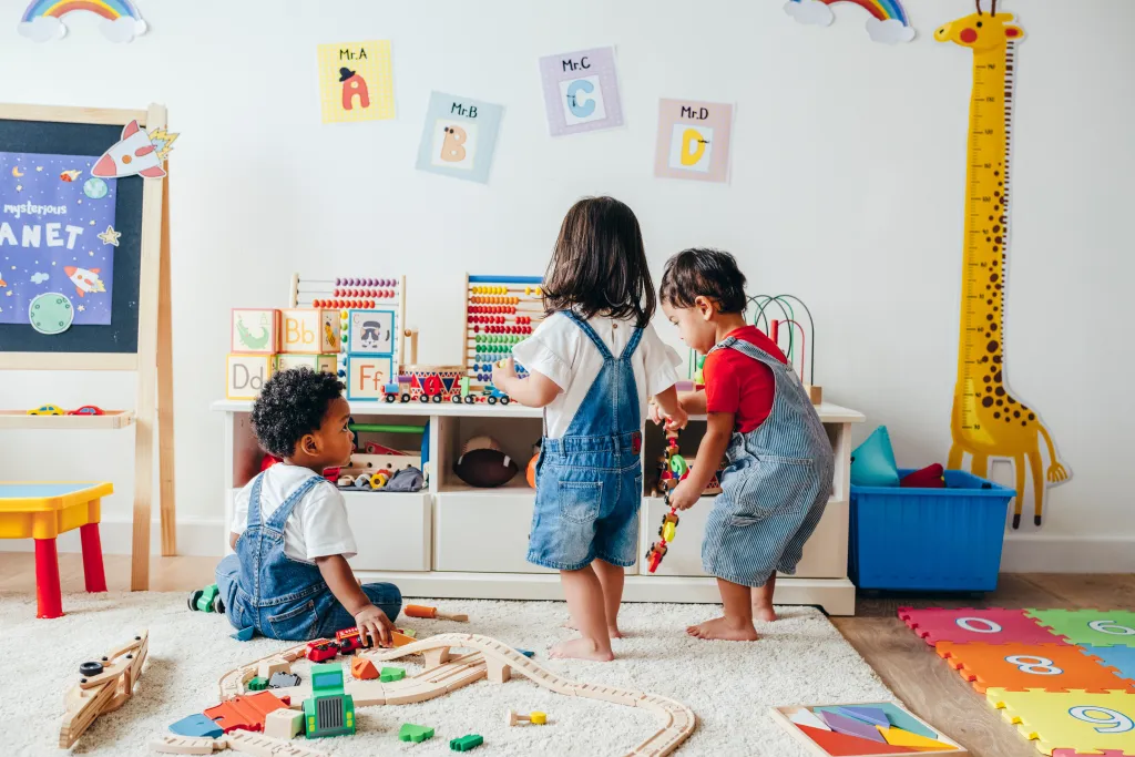 Three young children play with toys on a white rug in a brightly lit playroom.
