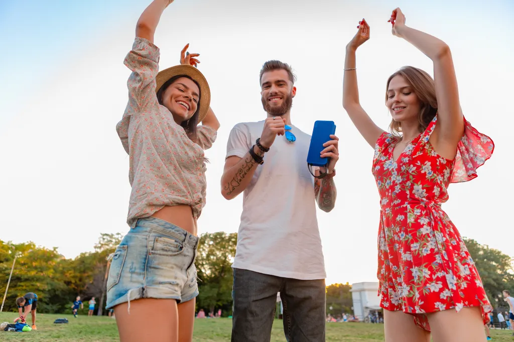 Three young friends dancing in a park, with a man holding a blue wireless speaker.