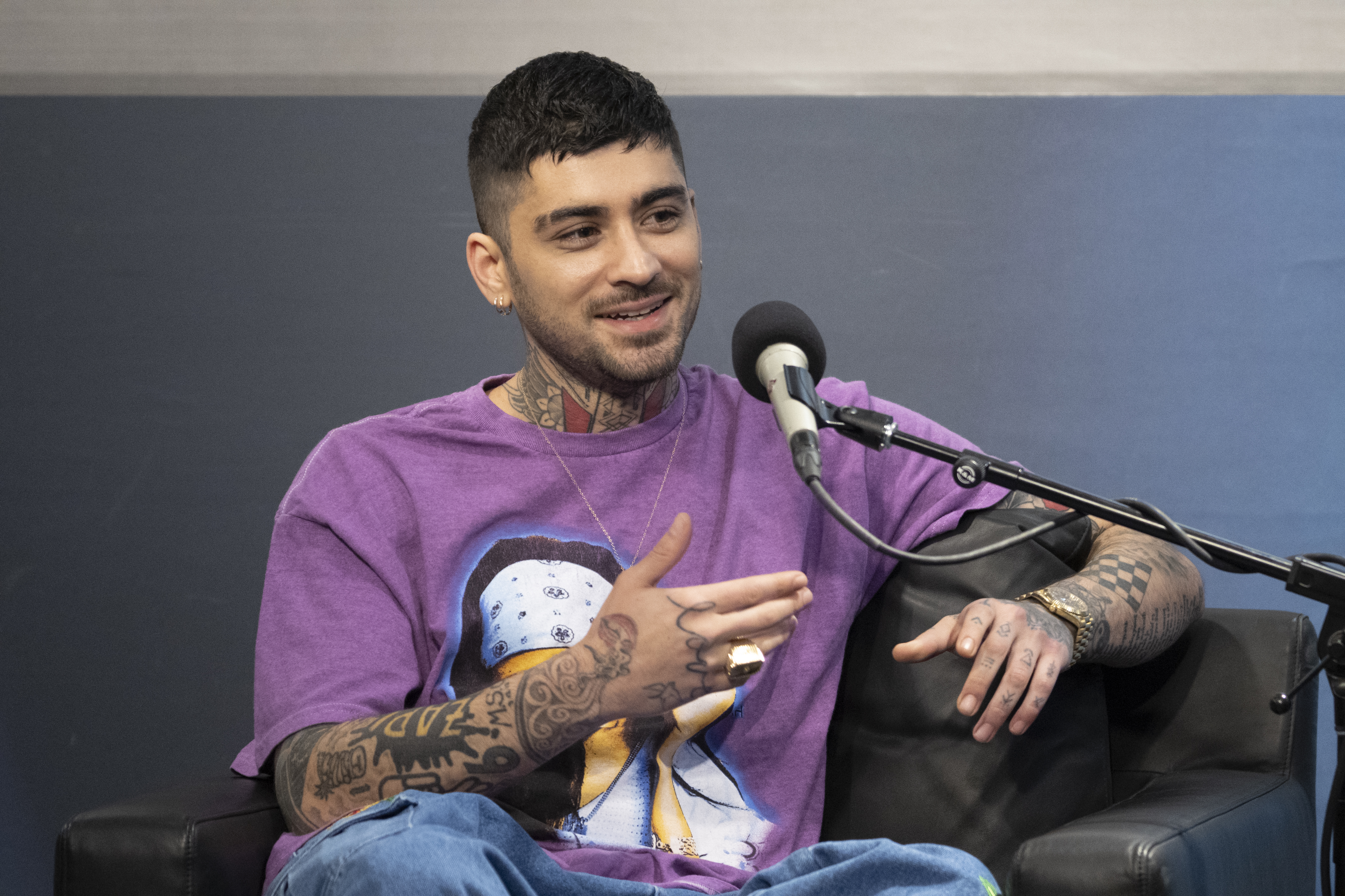 Zayn Malik, tattooed and wearing a purple shirt, smiles while speaking into a microphone.