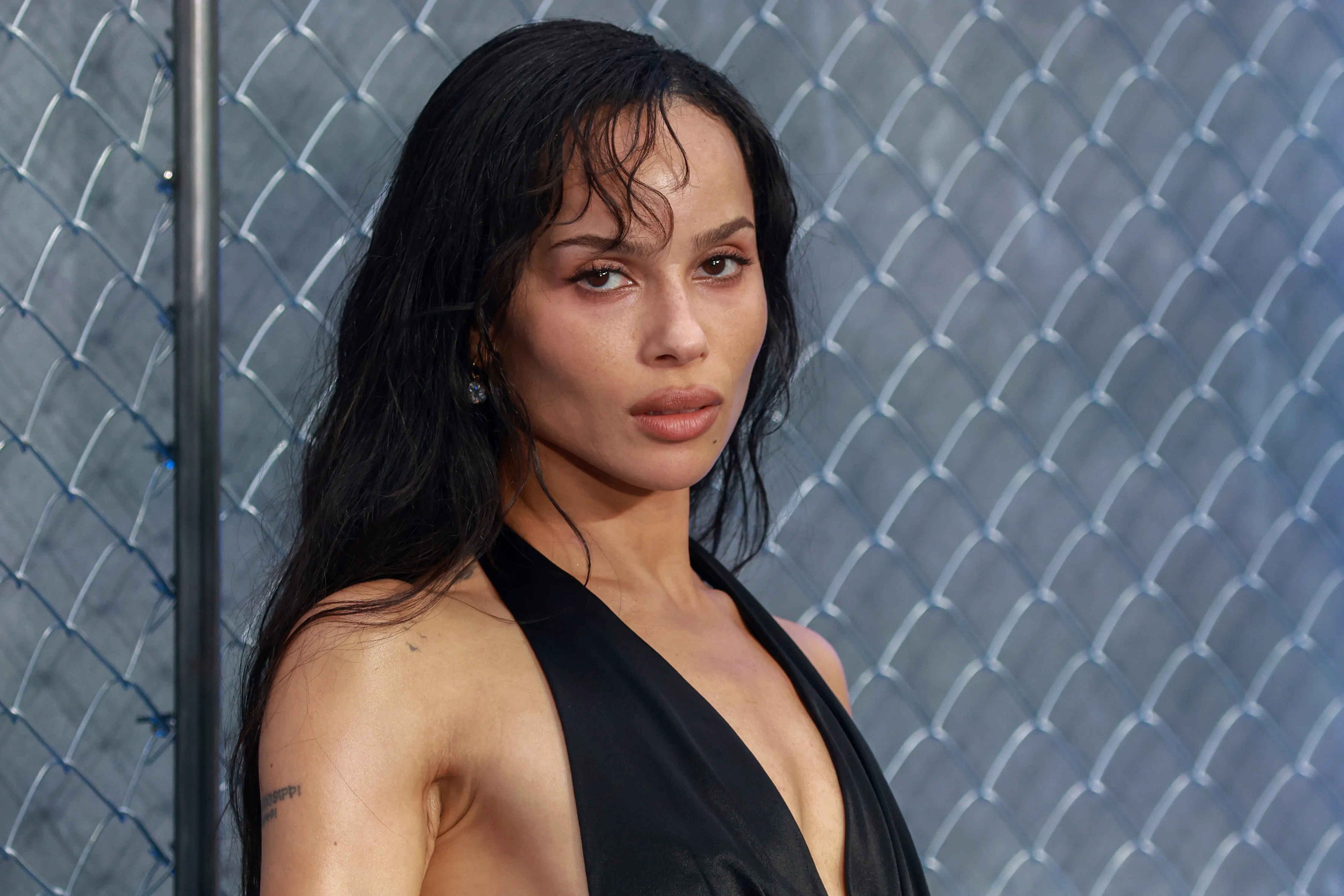 Zoë Kravitz with dark hair and a black top, against a fence background.