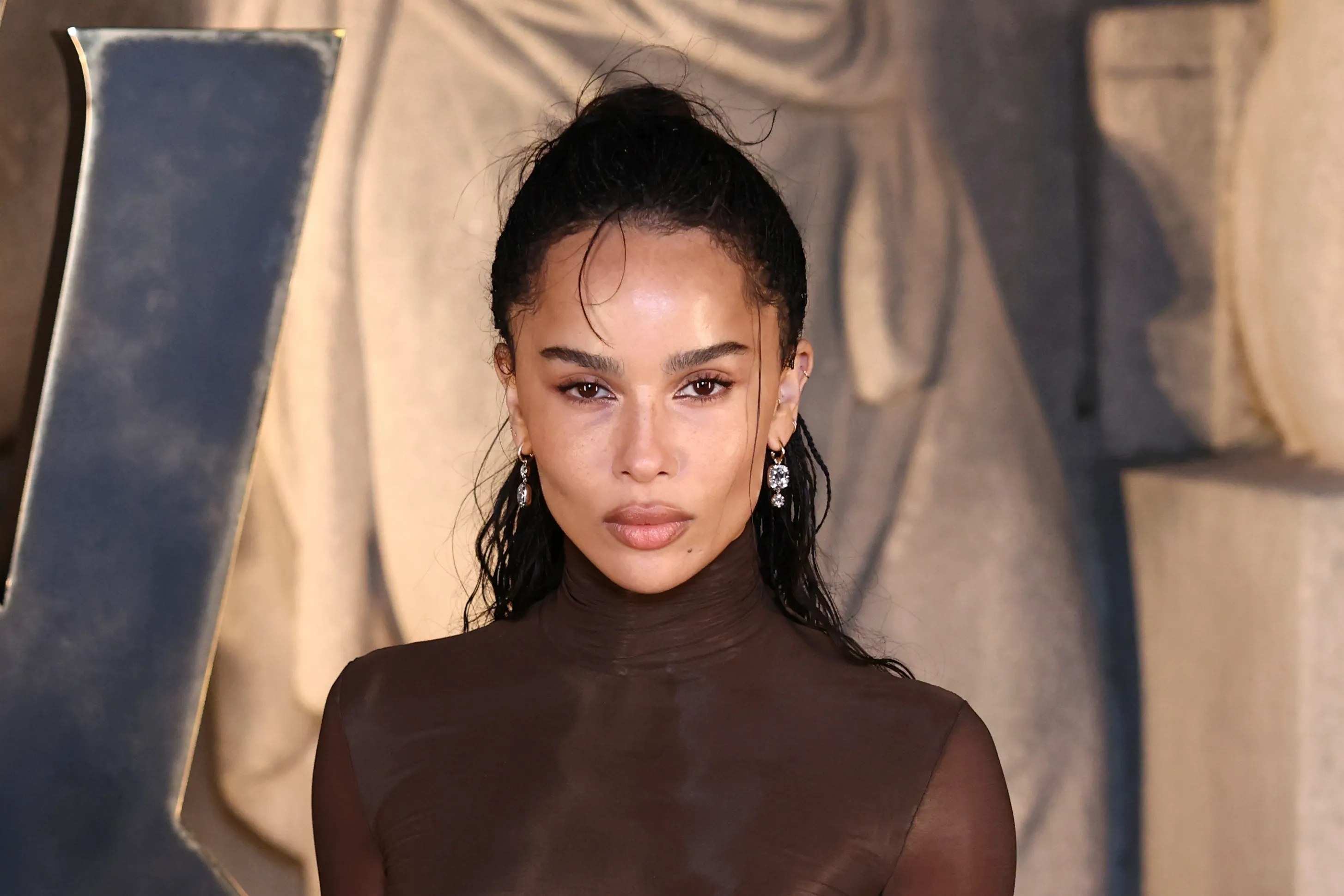 Zoe Kravitz with her hair pulled back, wearing a sheer brown top and chandelier earrings.