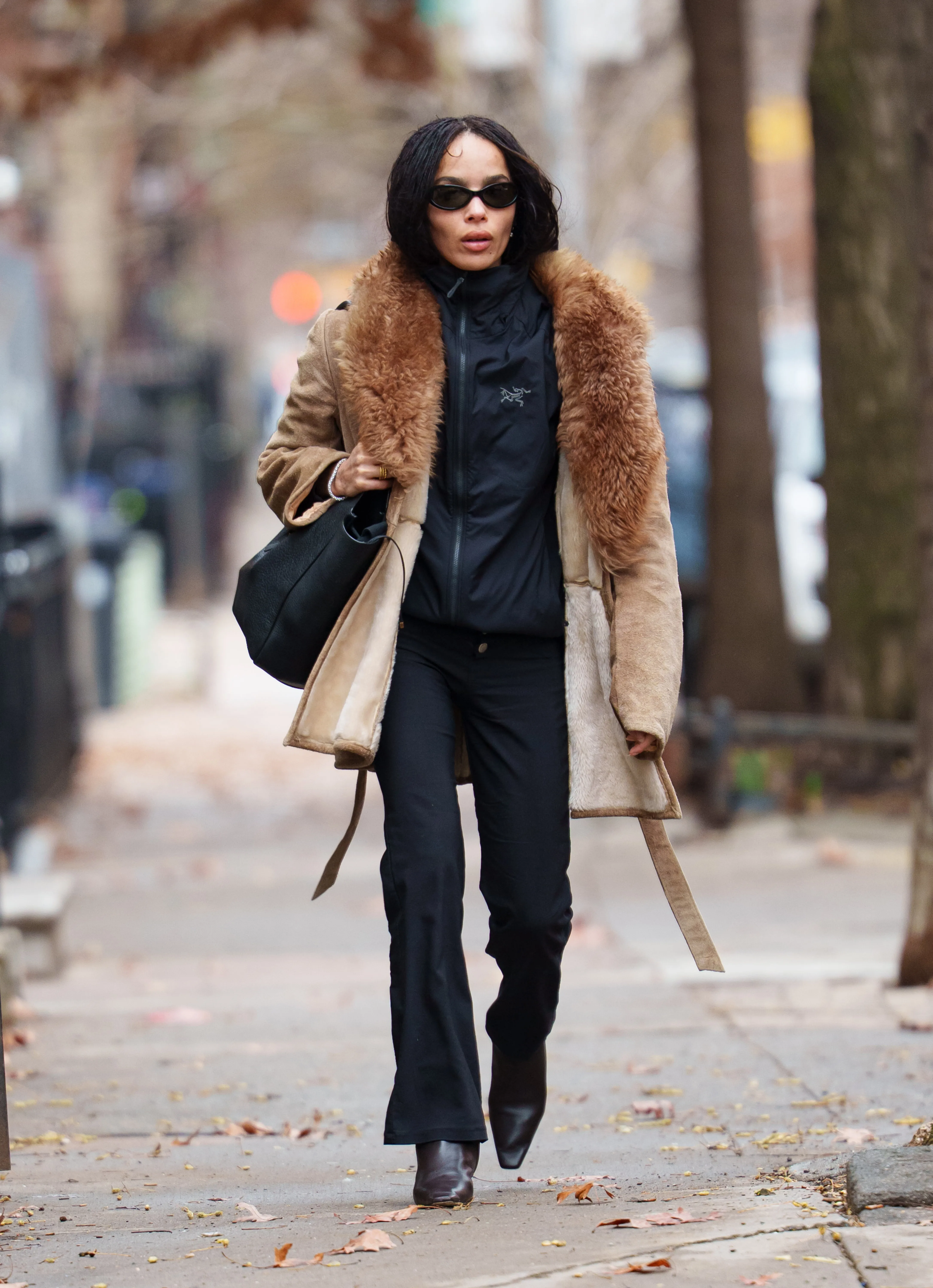 Zoe Kravitz walking outdoors in a tan and brown fur-lined coat, black top, and black pants.