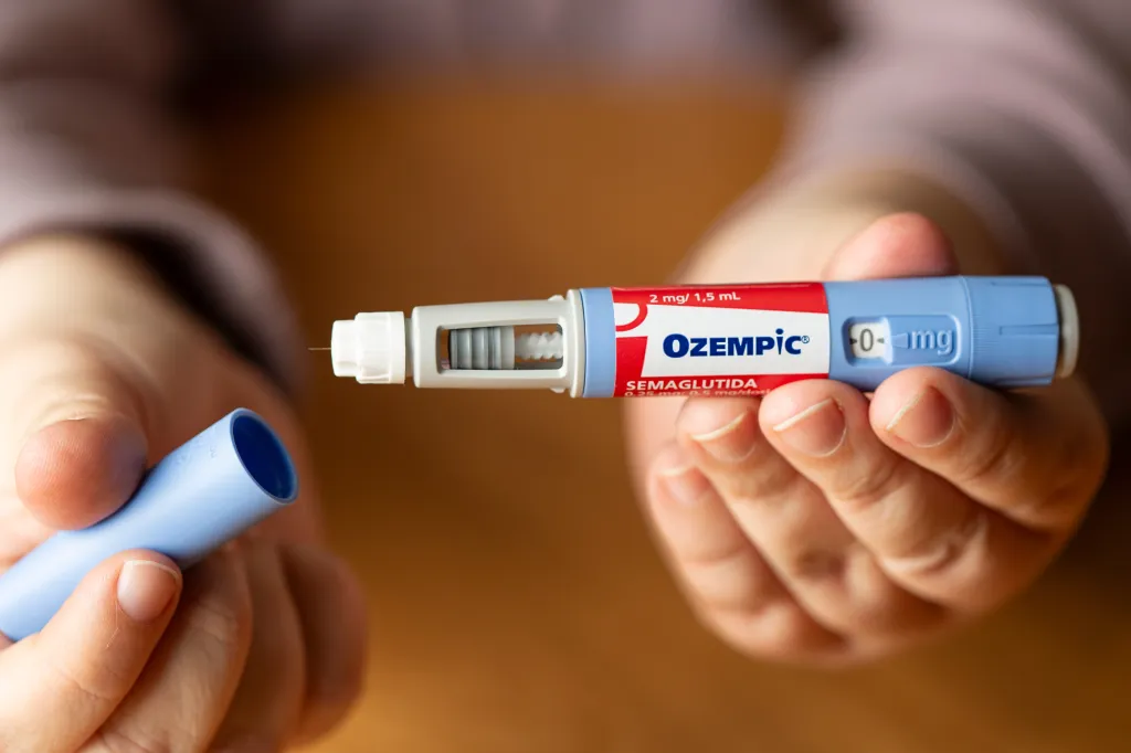 Hands holding an Ozempic semaglutide injection pen with the cap removed, revealing the needle