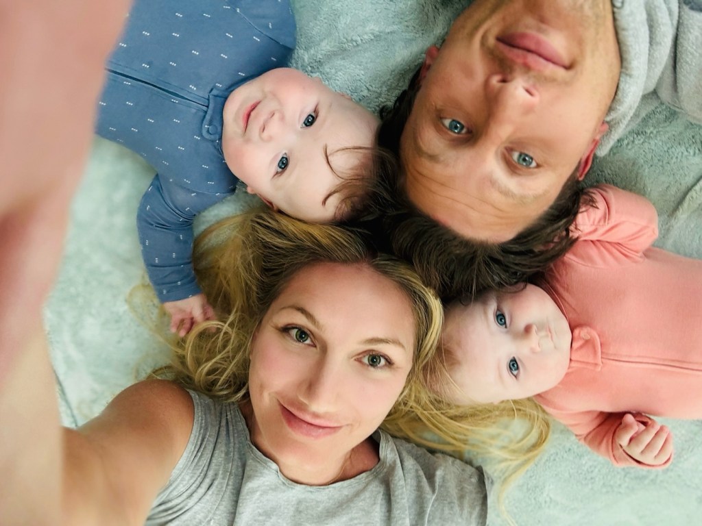 A family of four, including two adults and two babies, lying on a blanket and looking up at the camera.