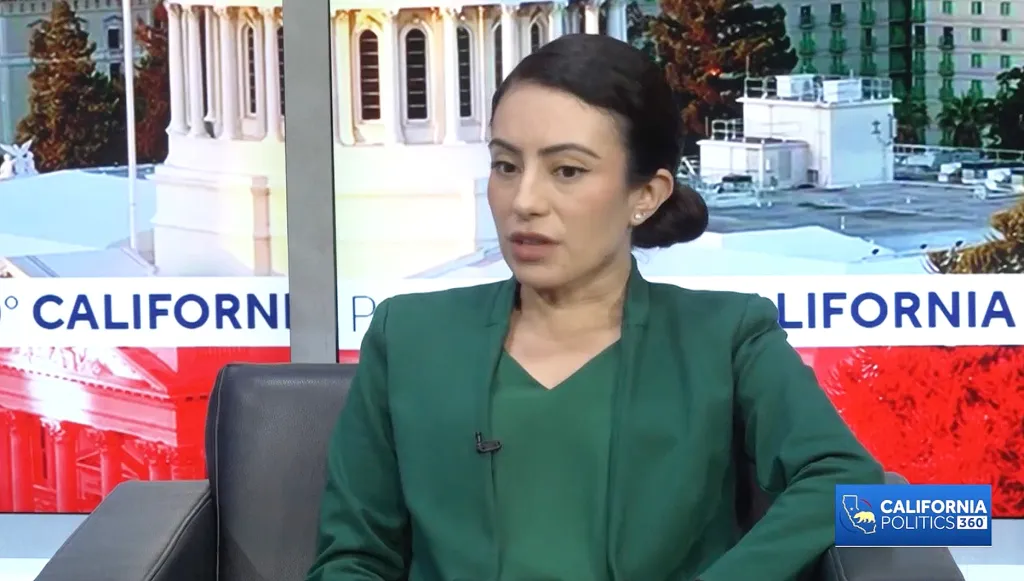A woman in a green jacket speaking on "California Politics 360" in front of a backdrop showing the California State Capitol building.