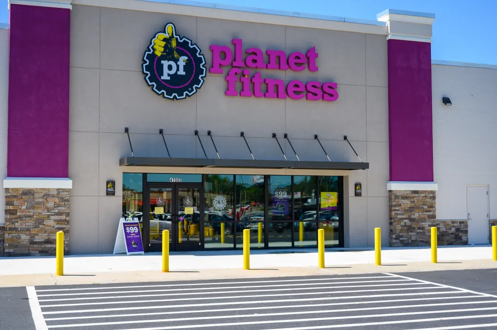 Exterior view of a Planet Fitness gym with purple and beige walls, glass doors, and a sign advertising a "$99 one year, one payment" special.