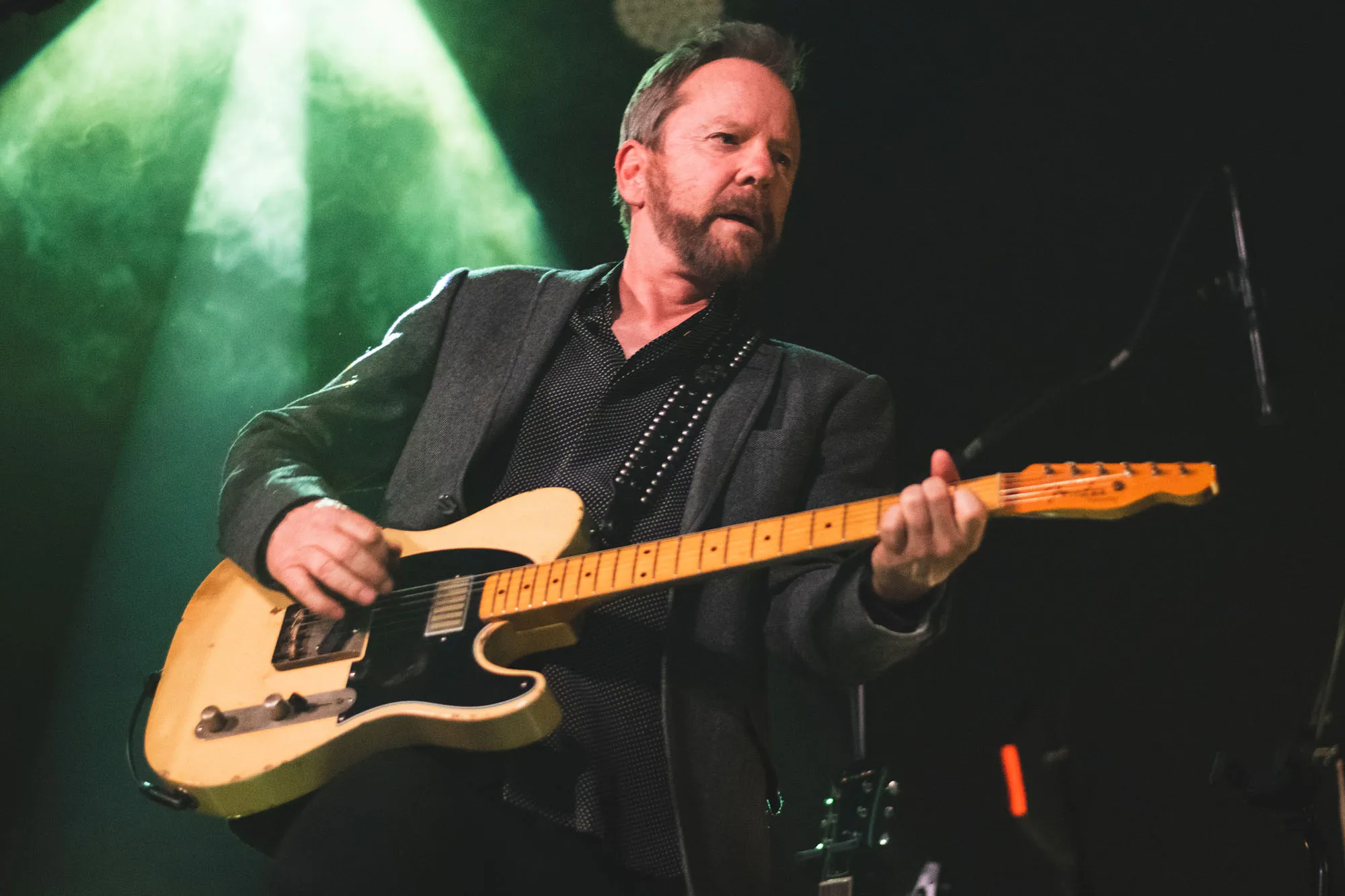 Kiefer Sutherland playing an electric guitar on stage under green stage lights.