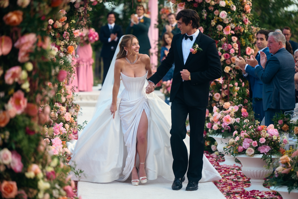 Bride and groom walk down an aisle surrounded by flowers and onlookers clapping.