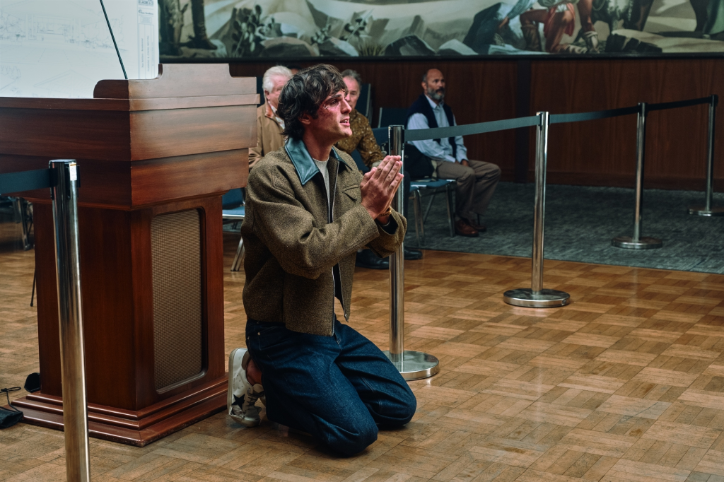 A man with an injured face kneels, hands pressed together, in a room with a large wooden podium and people seated in the background.