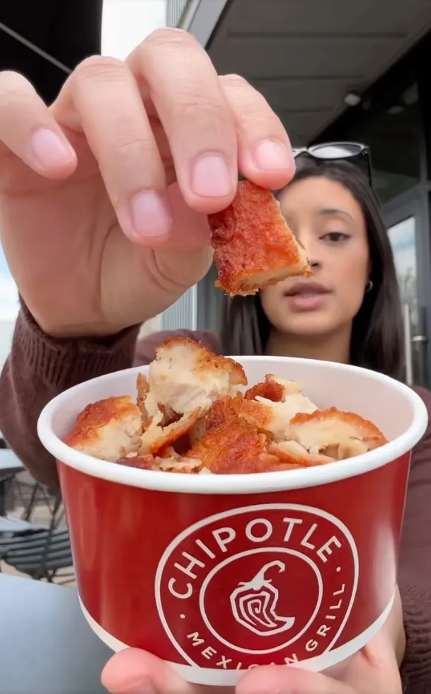 A person holds a piece of crispy chicken above a Chipotle cup full of more crispy chicken.
