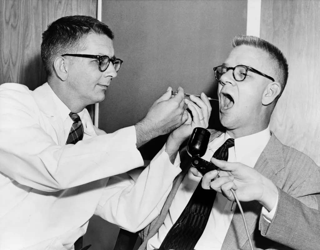 Dr. Harry L. Williams administering LSD to Carl Curt Pfeiffer, who has his mouth open while holding a microphone.