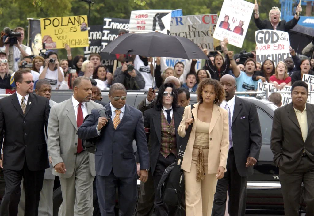 Michael Jackson, wearing sunglasses and a patterned vest, walks through a crowd of fans holding "Poland Loves You Michael" and "God is on Your Side Michael" signs, with bodyguards and other individuals around him.