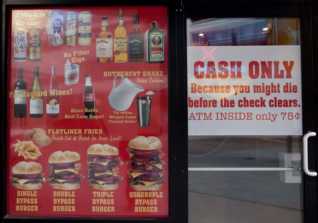 Menu and warnings posted in the window of the Heart Attack Grill in Las Vegas, showing items like "Bypass" burgers and "Flatliner" fries, and a "CASH ONLY" sign.