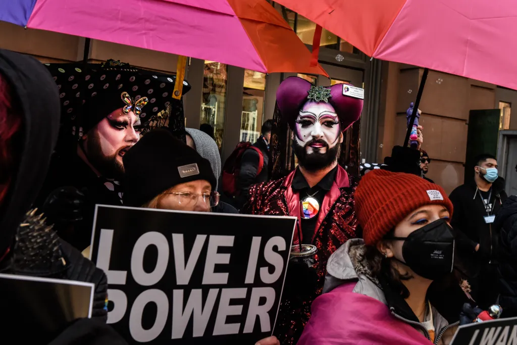 Protesters with signs, including one that reads "LOVE IS POWER," gather under pink and red umbrellas, with some in drag makeup and costumes.