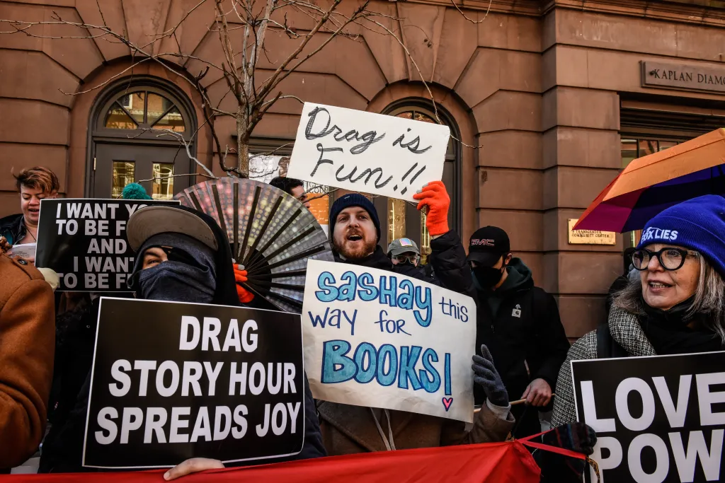 Protesters at a Drag Story Hour hold signs, one reads "Drag is Fun!" and another "Drag Story Hour Spreads Joy."