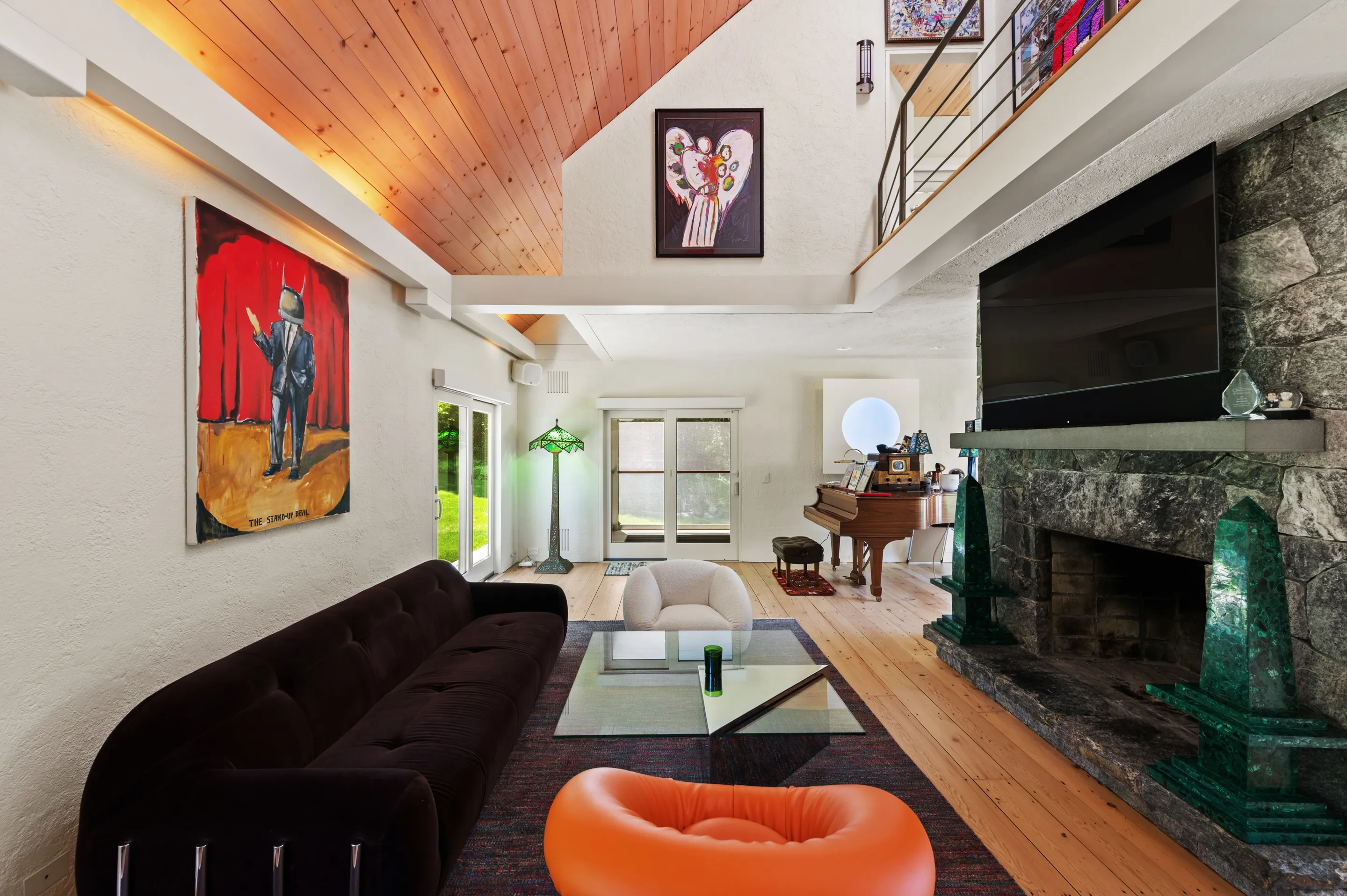 A spacious living room featuring a long, dark velvet sofa, a white armchair, and an orange pouf around a glass coffee table on a patterned rug.